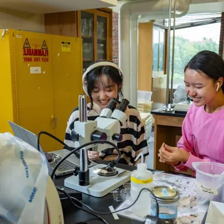 Two students, one in pink and one in stripes with headphones and earbuds, work at microscopes at Professor Marney Pratt's lab in Sabin-Reed